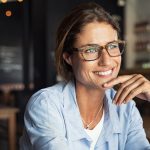 A woman wearing glasses and a light blue shirt smiles while resting her chin on her hand in a cafe setting.