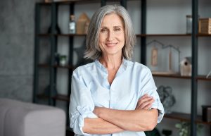 A woman with gray hair and a blue shirt stands smiling with arms crossed in front of a bookshelf.